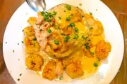 The image displays a plate of what appears to be a creamy seafood dish with shrimp and a garnish of chopped green onions, served on a bed of fried or perhaps grilled items that could be vegetables or a type of fritter.
