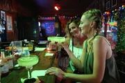 Two women are smiling and enjoying large margaritas at a festively decorated bar.