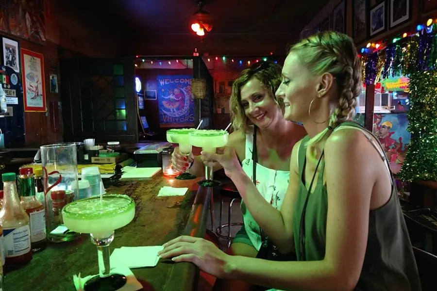 Two women are smiling and enjoying large margaritas at a festively decorated bar.