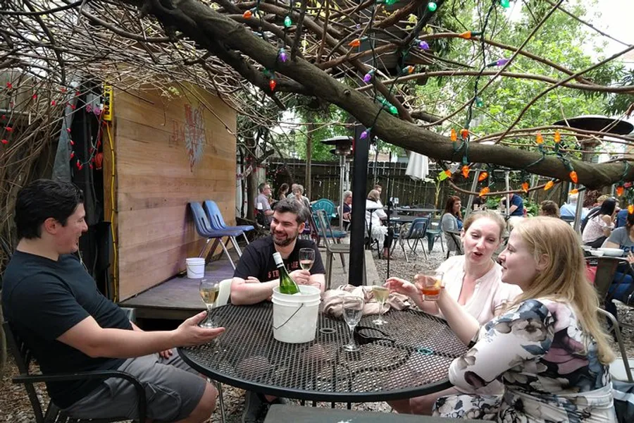 Four people are enjoying drinks and conversation at an outdoor table adorned with string lights, set beneath a rustic arbor.