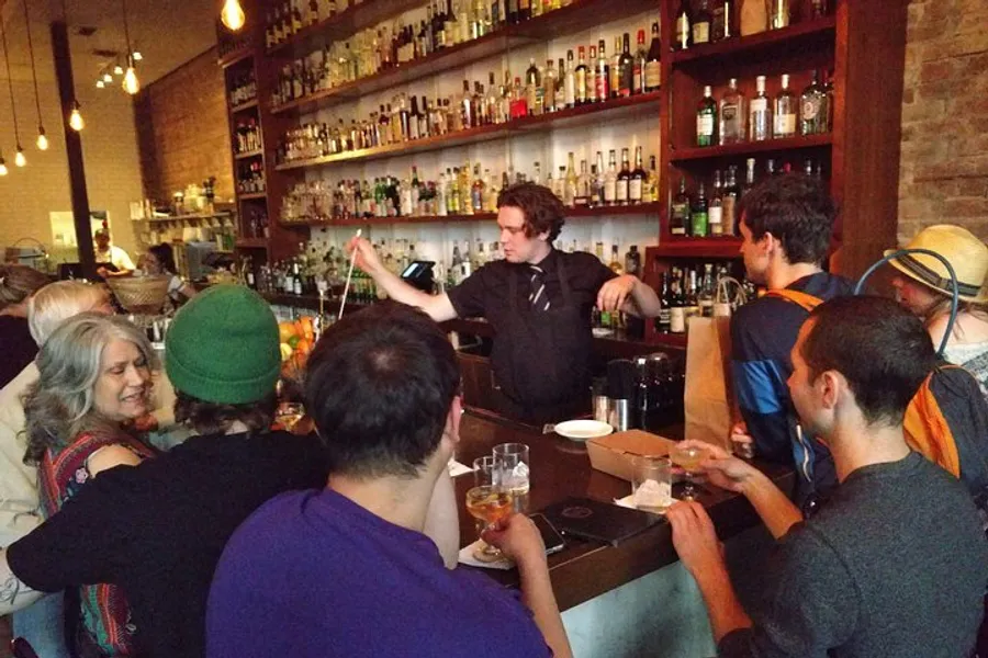 A bartender is pouring a drink behind a crowded bar with patrons engaged in conversation and holding beverages.