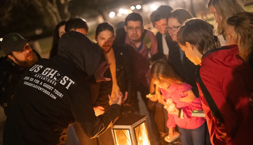 A group of people gather around a lantern at night seemingly engaged in a ghost tour led by a guide whose jacket advertises the adventure