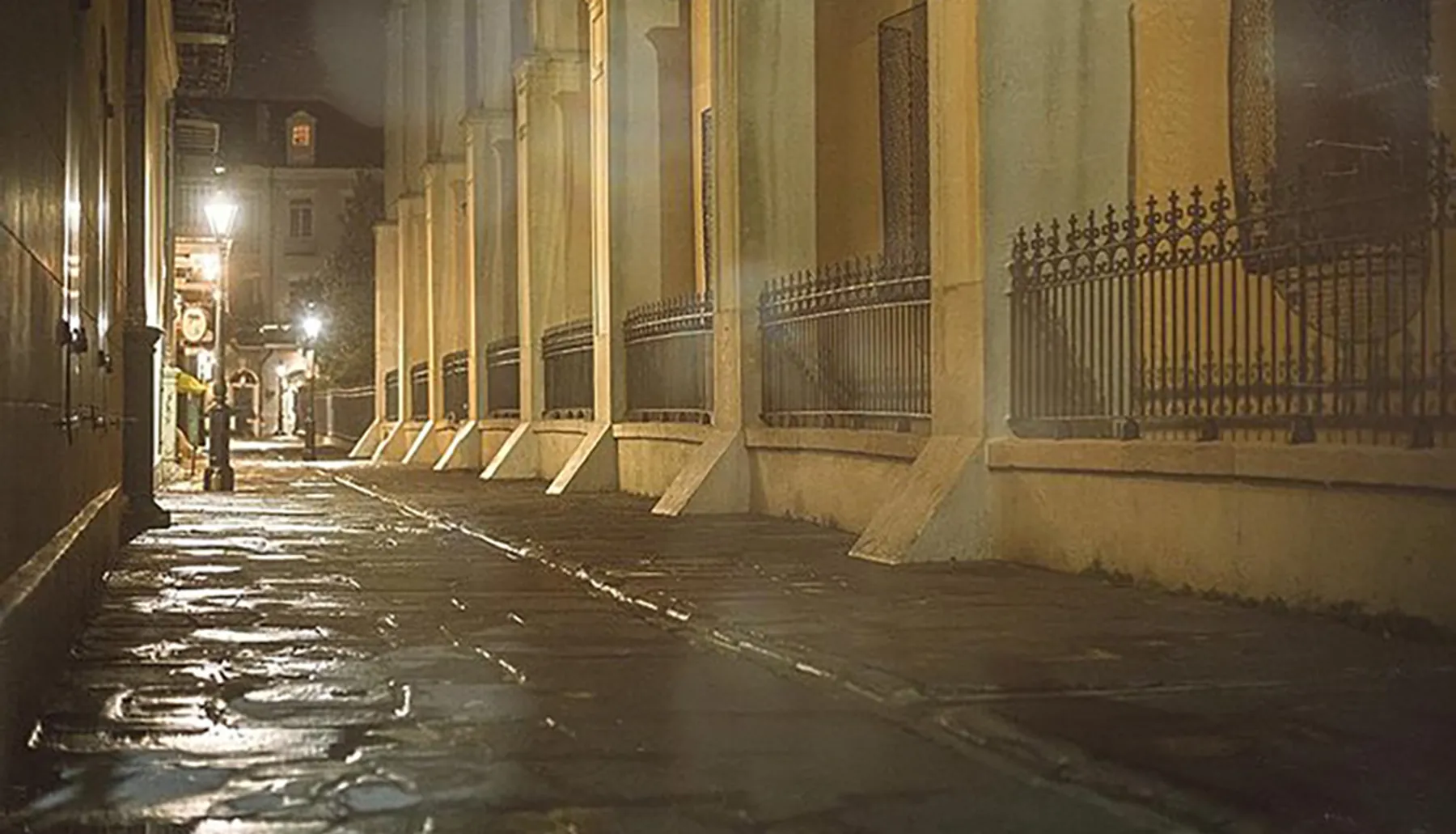 A dimly lit, wet cobblestone street flanked by stately buildings and wrought iron fences conveys a serene, mysterious atmosphere at night.