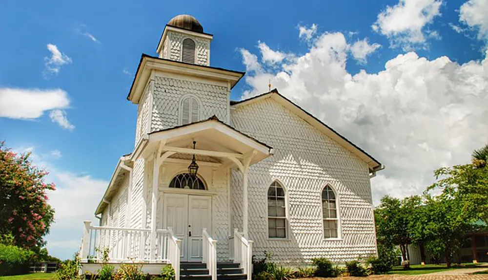 The image depicts a quaint white church with decorative wood shingles and a small bell tower against a backdrop of a blue sky dotted with fluffy clouds
