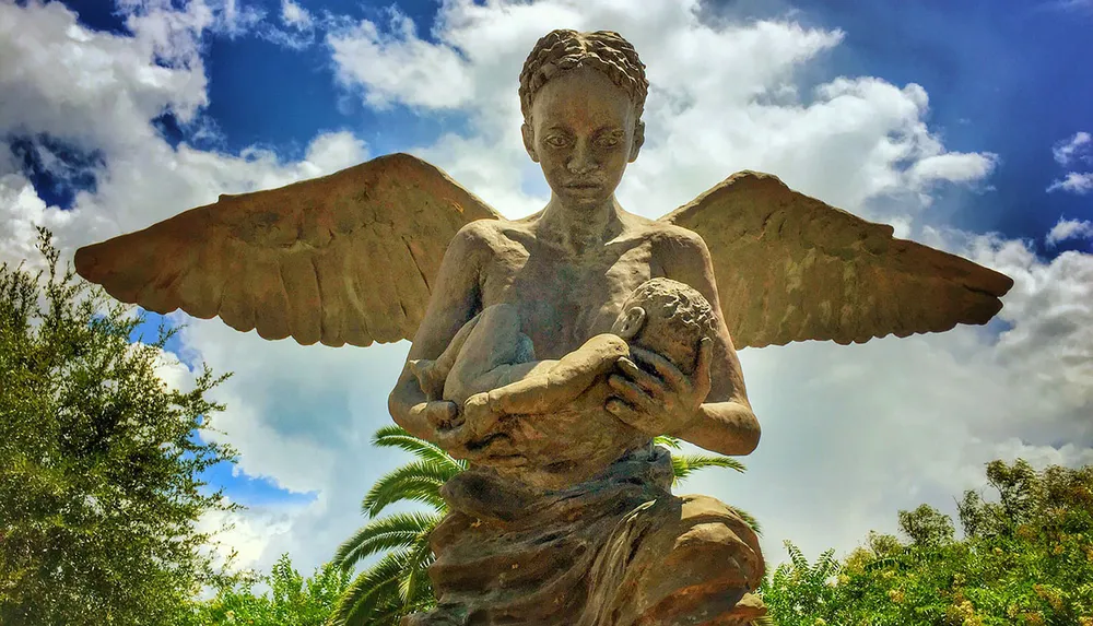 The image shows a weathered statue of an angelic figure with outstretched wings cradling a spherical object set against a cloudy sky and green foliage