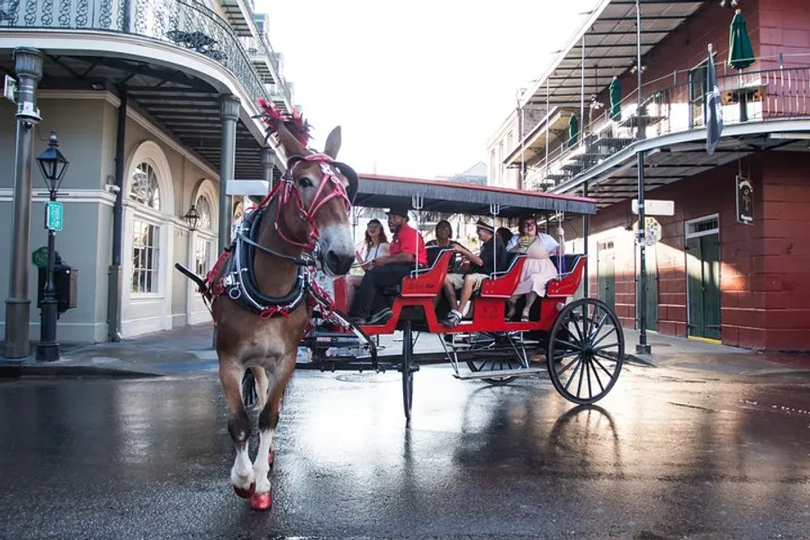 A horse pulling a red carriage with passengers on a sunny day through a street that hints at a New Orleans-style architecture.