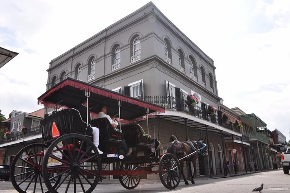 A horse-drawn carriage with passengers is parked in front of a classic two-story building with wrought iron balconies in what looks like a historical urban setting