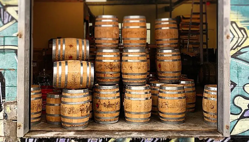 The image shows a collection of stacked wooden barrels possibly used for aging spirits or wine with a graffiti-painted wall as the background