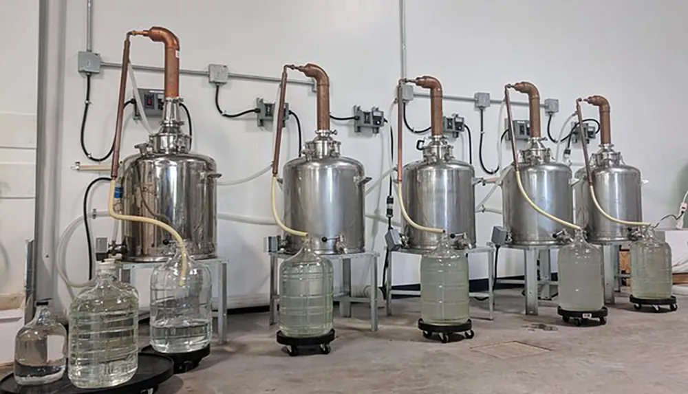 The image shows a row of distillation apparatuses possibly for chemical processing or the distillation of spirits with large stainless steel boilers connected to condensing columns and glass collection vessels