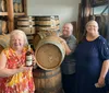 Three smiling women are posing in a room filled with wooden barrels likely at a distillery or winery with one woman holding a bottle of what appears to be liquor