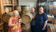 Three smiling women are posing in a room filled with wooden barrels, likely at a distillery or winery, with one woman holding a bottle of what appears to be liquor.