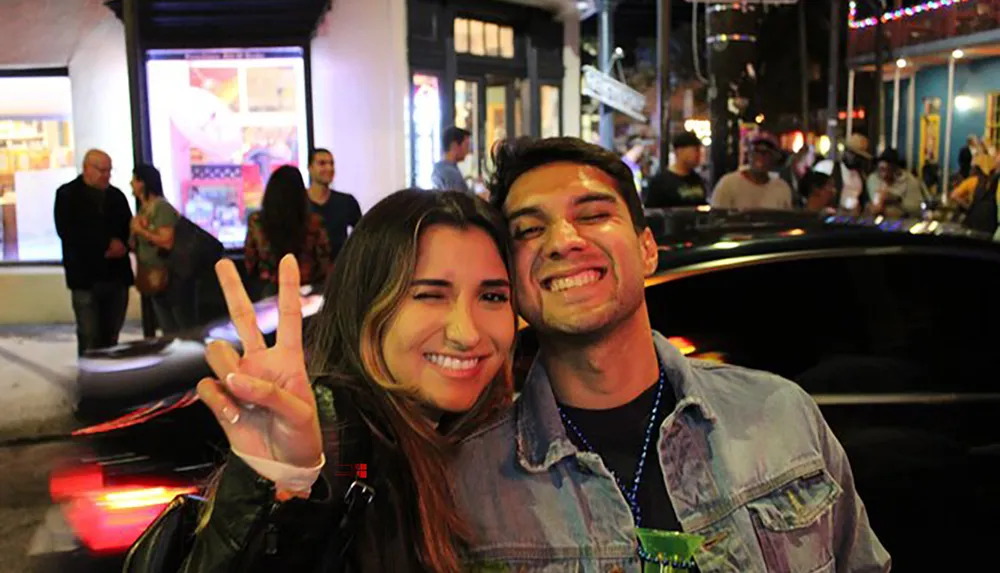 A smiling man and woman pose for a selfie on a lively urban street at night with the woman making a peace sign