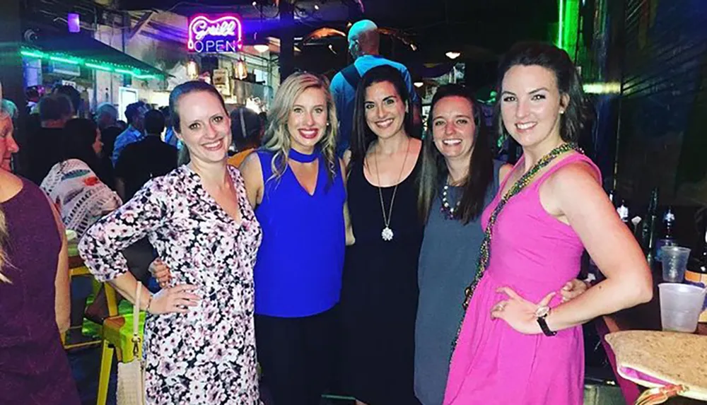 Five smiling women are posing together for a photo in a lively bar setting with a neon Open sign in the background
