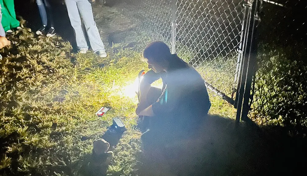 A person is sitting on the ground outdoors at night lit by a bright light with onlookers standing by a fence