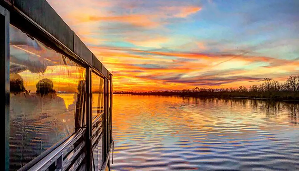 A vibrant sunset reflects on both the calm water of a lake and the glossy side of a boat creating a symmetrical display of warm colors and tranquility