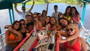 A group of young adults is enjoying a sunny day on a boat, posing with drinks and smiling for a group photo.