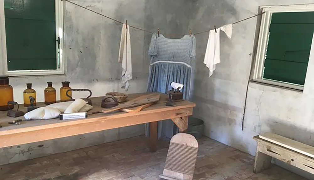 The image depicts a rustic room with an old-fashioned laundry setup including clothing hung to dry a washboard wooden ironing table vintage flatiron and brown glass bottles evoking a bygone era