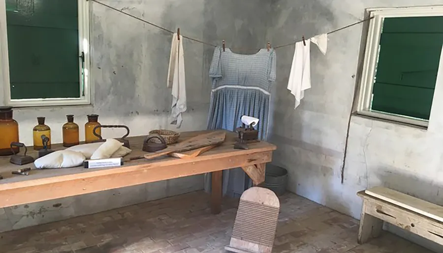 The image depicts a rustic room with an old-fashioned laundry setup, including clothing hung to dry, a washboard, wooden ironing table, vintage flatiron, and brown glass bottles, evoking a bygone era.