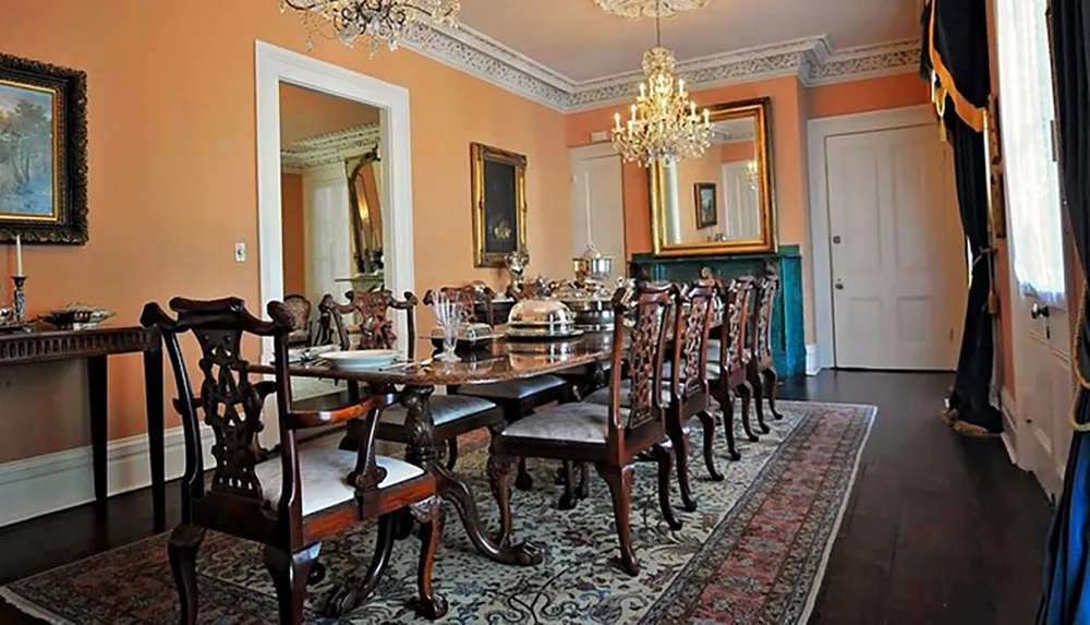 An elegant dining room with a long table set with silverware and glasses adorned by ornate chairs chandeliers and framed artwork