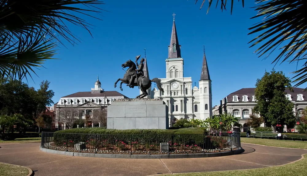 The image shows a statue of a mounted figure prominently displayed in front of a cathedral with two spires under a clear blue sky framed by palm leaves