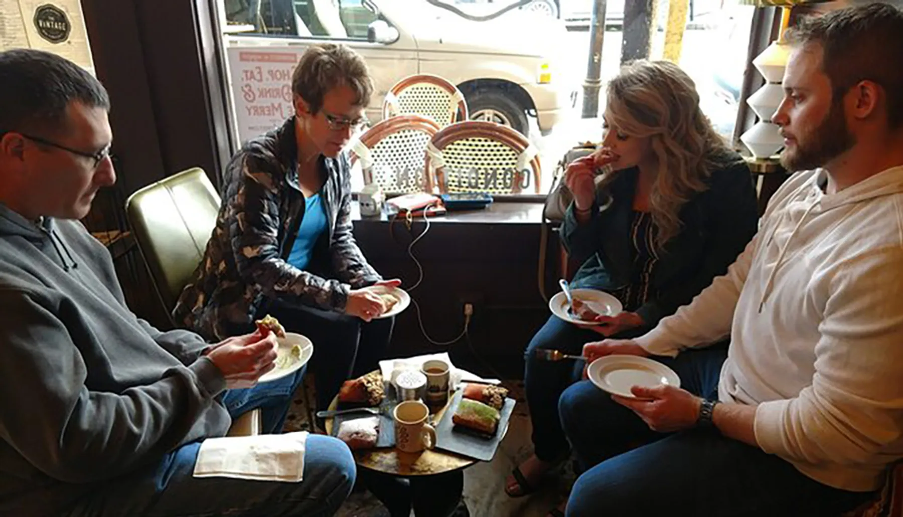 Four people are sitting in a cozy coffee shop setting, enjoying beverages and pastries while engaged in conversation.