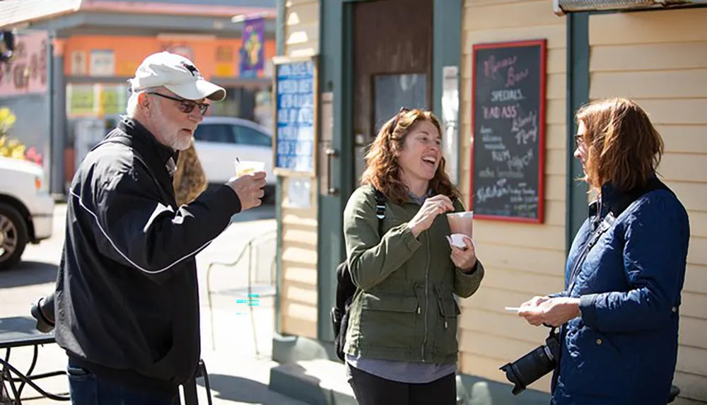 Three people are conversing and laughing on a sunny street while holding cups with a colorful storefront and a chalkboard with specials in the background