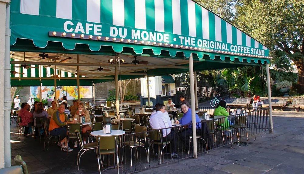 The image shows people seated at outdoor tables enjoying their time at Caf du Monde a renowned coffee stand known for its beignets in New Orleans