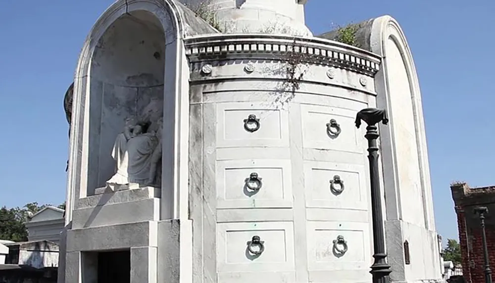 The image shows an ornate white mausoleum with sculptural elements and multiple niches set against a clear blue sky