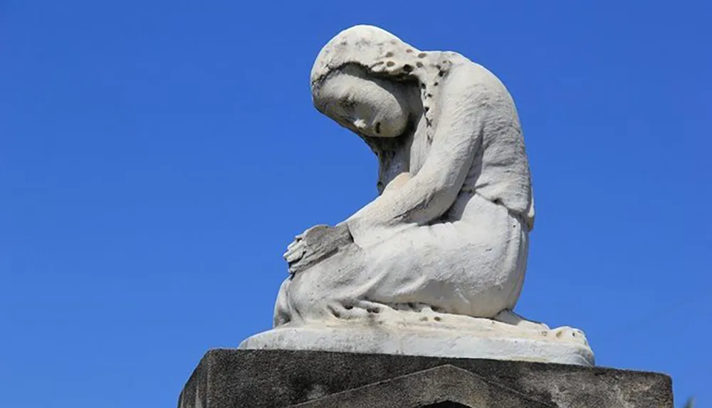 The image shows a weathered white stone sculpture of an anguished figure hunched over in a pose that suggests deep sorrow set against a clear blue sky