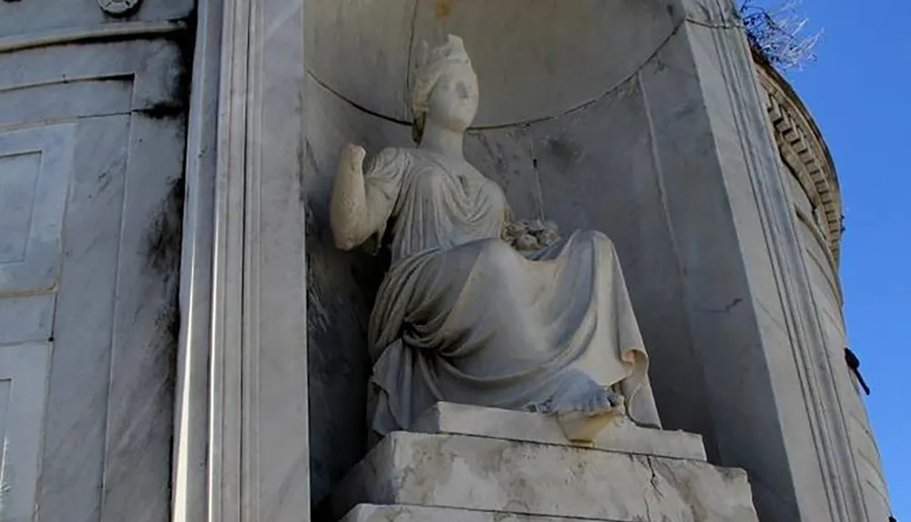 The image shows a classical statue of a robed figure with a headpiece sitting on a pedestal part of a historical monument or structure