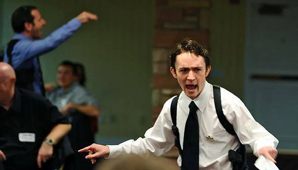 A man in a white shirt and suspenders is angrily gesturing and speaking while another man in the background points and seems to be shouting both in a room with other people who appear to be in a state of commotion