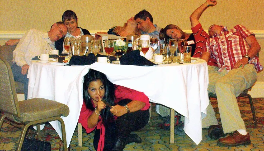 A woman is playfully gesturing silence while hiding under a table as others around her appear to be asleep in their chairs amongst a scattered array of dishes and glasses suggesting the aftermath of a lively event