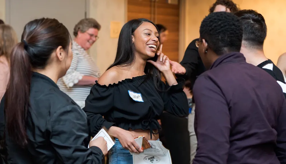 A group of people is engaging in conversation at what appears to be a social or networking event with one woman in the center smiling broadly and touching her chin creating a focal point for the image