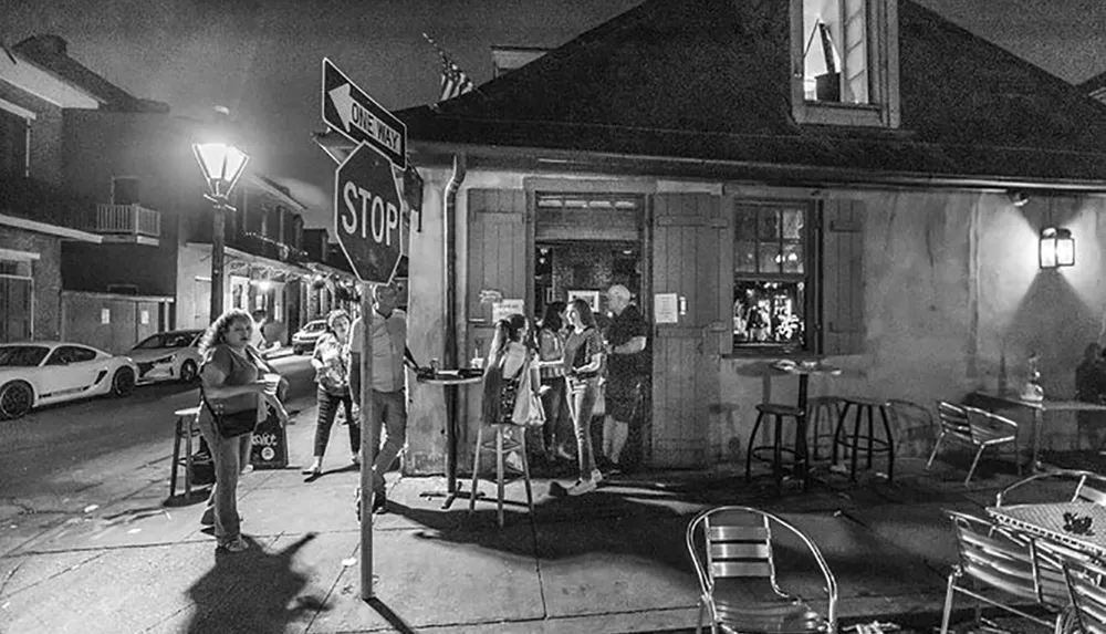 A black and white photo showing people gathering at a corner bar at night with a stop sign and street lamps illuminated