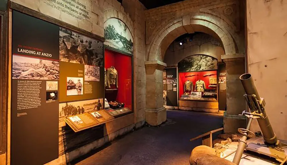 This image shows an exhibit in a museum dedicated to military history featuring historical photographs uniforms and artillery displayed in an evocative dimly-lit setting