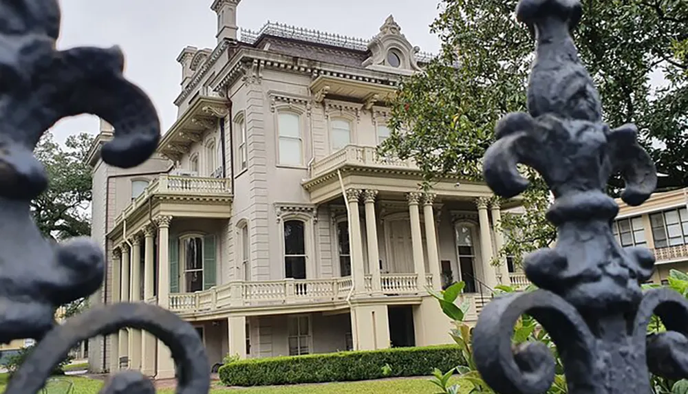An elegant historical mansion is viewed through an ornate black iron fence featuring classical architectural elements and a lush front garden