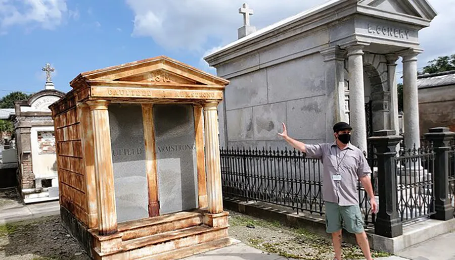 A person wearing a mask is gesturing towards an ornate mausoleum with the name Gaultier on it in a cemetery with above-ground tombs.