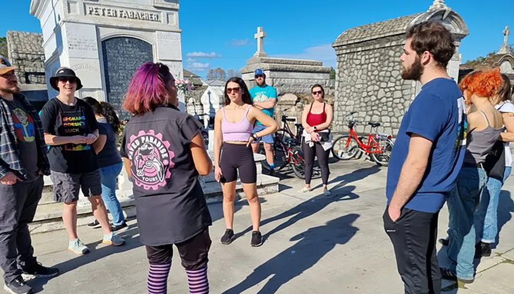 A group of people is attentively listening to a guide dressed in black and pink during a tour in a cemetery on a sunny day