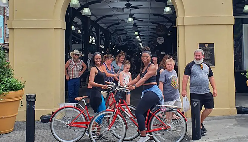 The image shows a lively street scene with two women smiling at the camera while sitting on red bicycles as various other people walk by in a covered market area