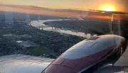 The image captures a stunning sunset view from an airplane window, showing the plane's engine, reflective wing, and a meandering river below as the sun dips towards the horizon.