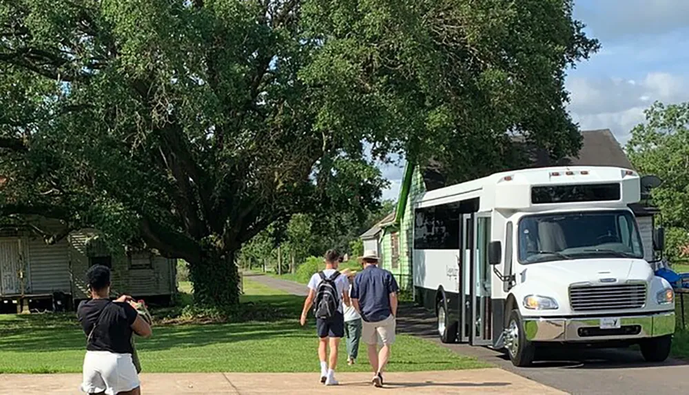 Two individuals are walking towards a white shuttle bus on a sunny day while another person stands in the foreground taking a photograph