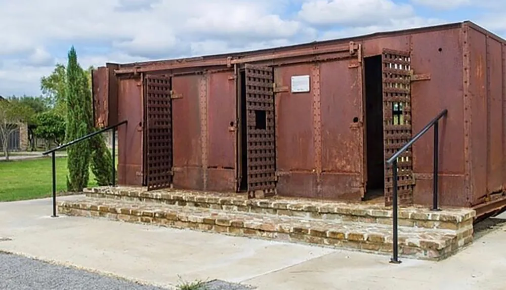 The image shows a large rusted iron structure with doors resembling a type of industrial equipment or containment unit mounted on a concrete foundation with steps leading up to it