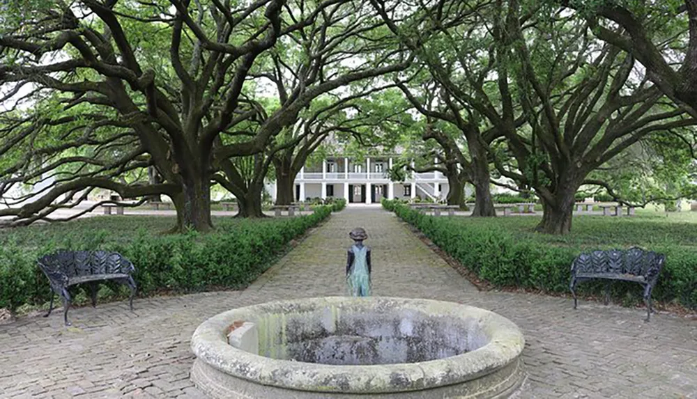 The image shows a pathway lined with majestic trees leading to an elegant white house with a small figure standing near a stone fountain in the foreground