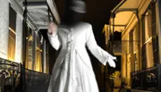 The image shows a blurred figure resembling a classic ghost, dressed in white with a blurred face, seemingly floating outside an illuminated window at night.