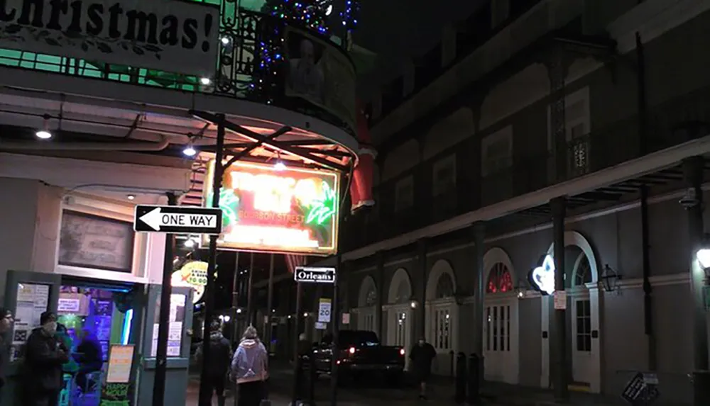 The image shows an urban street scene at night with neon signs a Christmas banner pedestrians and the balconied architecture typical of the French Quarter in New Orleans
