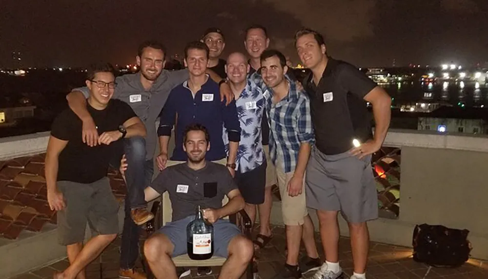 A group of smiling men pose for a photo at night on a rooftop with city lights in the background