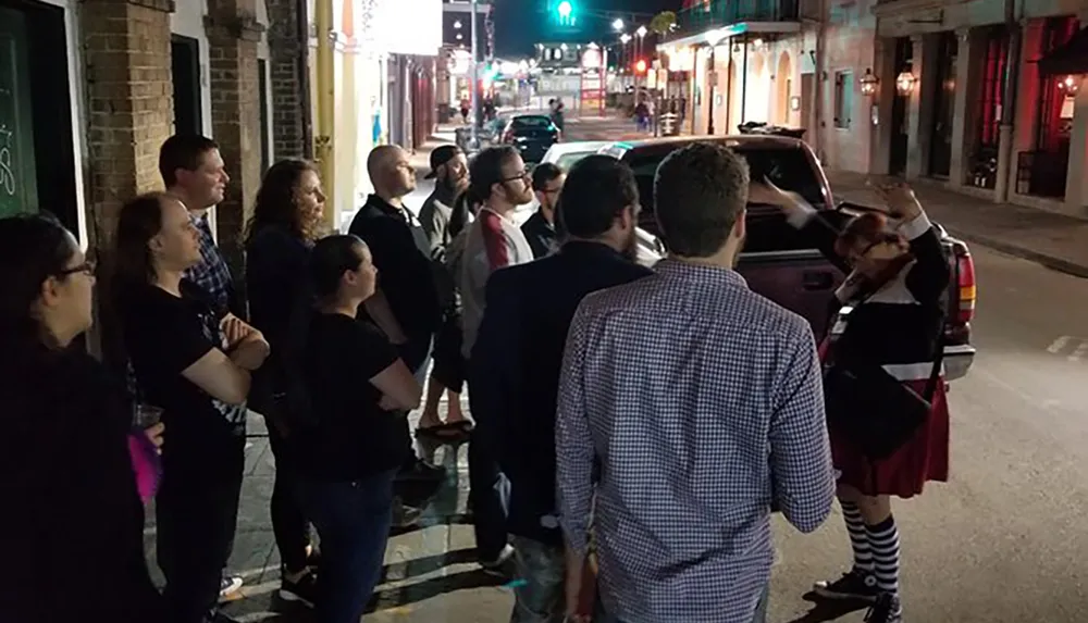 A group of people attentively listening to a person gesturing possibly a tour guide on a city street at night