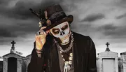 A person dressed in a suit with skull face paint tips their hat in a graveyard with a dramatic cloudy sky in the background.