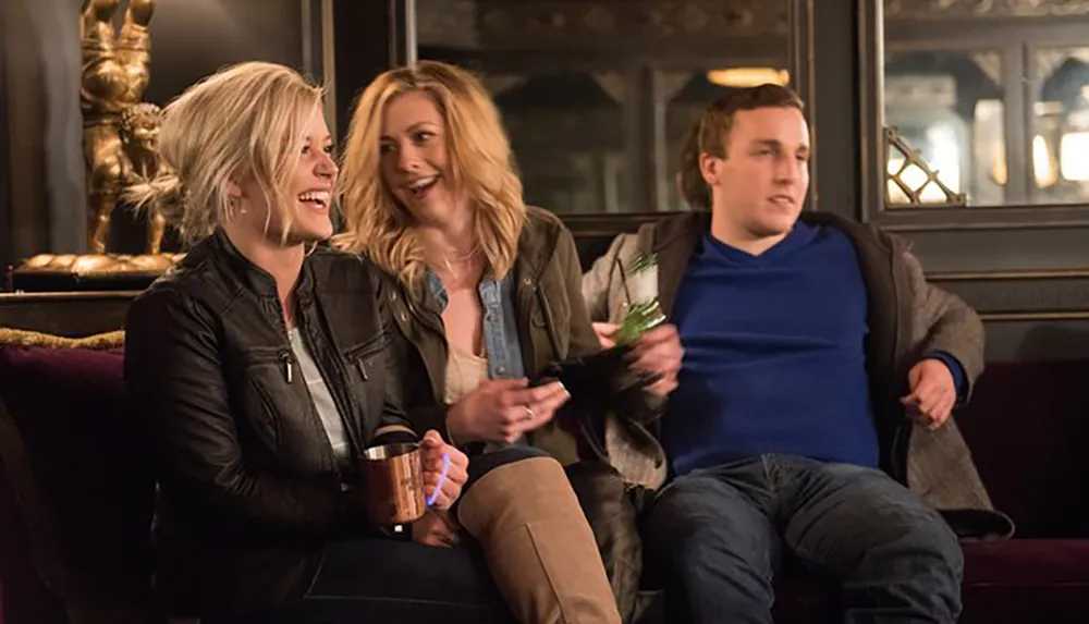 Three people are sitting on a couch with two of them laughing and having a good time while one looks disinterested and the setting appears to be a cozy indoor area possibly a bar or lounge
