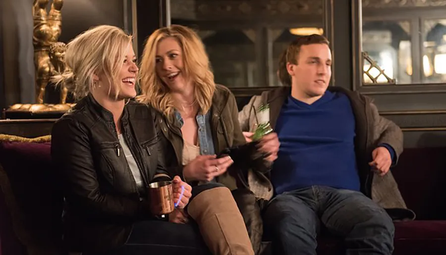 Three people are sitting on a couch with two of them laughing and having a good time while one looks disinterested, and the setting appears to be a cozy indoor area, possibly a bar or lounge.